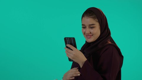 تصفح مواقع الشراء للاطفال بتعابير السرور، مفهوم الحمل والانجاب، الاهتمام بصحة الجنين، الحفاظ على صحتها و صحة طفلها، الاستفادة من العروض الموسمية والتخفيضات، تصوير مقرب لإمراة عربية خليجة سعودية ترتدي العباءة والحجاب تحمل أكياس تسوق ملونة، خلفية زرقاء