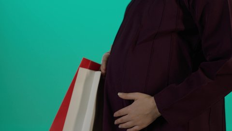 شراء حاجات الطفل الأول بتعابير السرور، مفهوم الحمل والانجاب، الاهتمام بصحة الجنين، الحفاظ على صحتها و صحة طفلها، الاستفادة من العروض الموسمية والتخفيضات، تصوير مقرب لإمراة عربية خليجة سعودية ترتدي العباءة والحجاب تحمل أكياس تسوق ملونة، خلفية زرقاء