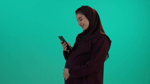 التثقف بالمعلومات عن الحمل على الهاتف المحمول، مفهوم الحمل والانجاب، الاهتمام بصحة الجنين، الحفاظ على صحتها و صحة طفلها، الاستفادة من العروض الموسمية والتخفيضات، تصوير مقرب لإمراة عربية خليجة سعودية ترتدي العباءة والحجاب تحمل أكياس تسوق ملونة وهاتف محمول، خلفية زرقاء