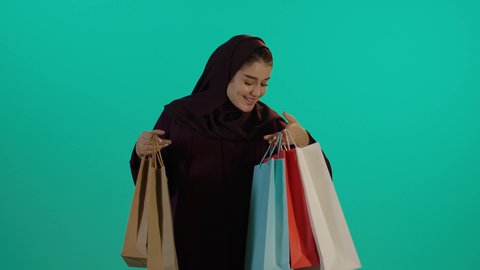 النظر الى اكياس التسوق خلال الحمل، مفهوم الحمل والانجاب، الاهتمام بصحة الجنين، الحفاظ على صحتها و صحة طفلها، الاستفادة من العروض الموسمية والتخفيضات، تصوير مقرب لإمراة عربية خليجة سعودية ترتدي العباءة والحجاب تحمل أكياس تسوق ملونة وهاتف محمول، خلفية زرقاء