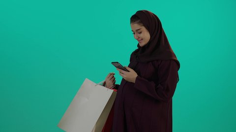 التسوق اونلاين خلال الحمل، مفهوم الحمل والانجاب، الاهتمام بصحة الجنين، الحفاظ على صحتها و صحة طفلها، الاستفادة من العروض الموسمية والتخفيضات، تصوير مقرب لإمراة عربية خليجة سعودية ترتدي العباءة والحجاب تحمل أكياس تسوق ملونة وهاتف محمول، خلفية زرقاء