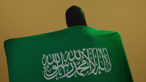 السعادة في إحياء ذكرى اليوم الوطني،  إظهار الاخلاص والحب للبلاد، الاحتفال بذكرى اليوم الوطني السعودي، يوم العلم السعودي 11 مارس، مفهوم حب الوطن والنتماء إليه،  بورتريه لامرأة عربية خليجية سعودية محجبة تقف وتضع علم الوطن على اكتافها بثقة واعتزاز، خلفية بيج.