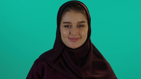 الحفاظ على صحتها و صحة طفلها، استخدام اصبع الابهام والقبول، مفهوم الحمل والانجاب، الاهتمام بصحة الجنين، إمرأة عربية خليجية سعودية تمسك بيدها حذاء أطفال، تصوير مقرب لإمراة عربية خليجة سعودية ترتدي العباءة والحجاب تنظر إلى الكاميرا بإيماءات السرور، خلفية زرقاء