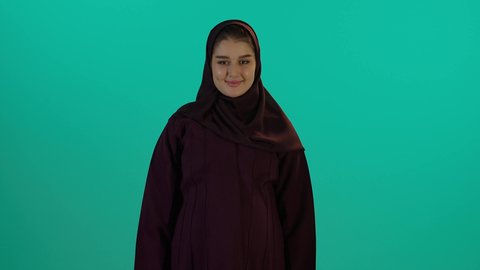 استخدام اصبع الابهام والقبول، مفهوم الحمل والانجاب، الاهتمام بصحة الجنين، الحفاظ على صحتها و صحة طفلها، إمرأة عربية خليجية سعودية تمسك بيدها حذاء أطفال، تصوير مقرب لإمراة عربية خليجة سعودية ترتدي العباءة والحجاب تنظر إلى الكاميرا بإيماءات السرور، خلفية زرقاء