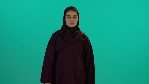 الاهتمام بصحة الجنين، مفهوم الحمل والانجاب، خفض الابهام للاسفل وعدم القبول، الحفاظ على صحتها و صحة طفلها، إمرأة عربية خليجية سعودية تمسك بيدها حذاء أطفال، تصوير مقرب لإمراة عربية خليجة سعودية ترتدي العباءة والحجاب تنظر إلى الكاميرا بإيماءات الغضب، خلفية زرقاء