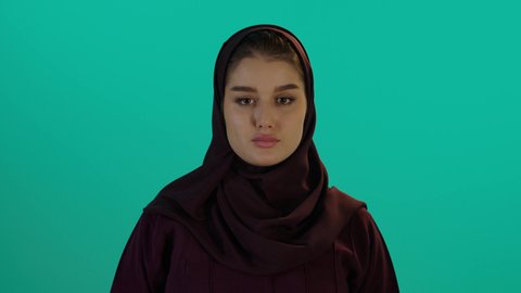 مفهوم الحمل والانجاب،خفض الابهام للاسفل وعدم القبول، الاهتمام بصحة الجنين، الحفاظ على صحتها و صحة طفلها، إمرأة عربية خليجية سعودية تمسك بيدها حذاء أطفال، تصوير مقرب لإمراة عربية خليجة سعودية ترتدي العباءة والحجاب تنظر إلى الكاميرا بإيماءات الغضب، خلفية زرقاء