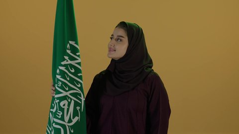 العيد الوطني السعودي 23 سبتمبر، إظهار الاخلاص والحب للبلاد، الاحتفال بذكرى اليوم الوطني السعودي، يوم العلم السعودي 11 مارس، مفهوم حب الوطن والنتماء إليه،  بورتريه لامرأة عربية خليجية سعودية محجبة تقف وتحمل علم الوطن بثقة واعتزاز، خلفية بيج.