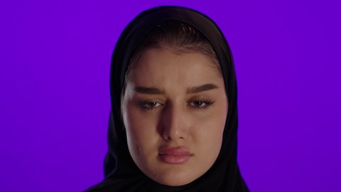 Anger and the demand to stop; the concept of pregnancy and childbirth; concern for the health of the fetus; maintaining her health and the health of her child; a Saudi Arabian woman from the Gulf holding a child's shoe; a close-up of a Saudi Arabian woman from the Gulf wearing an abaya and hijab, looking at the camera with thoughtful gestures; purple background.