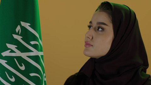 إظهار الاخلاص والحب للبلاد، الاحتفال بذكرى اليوم الوطني السعودي، يوم العلم السعودي 11 مارس، مفهوم حب الوطن والنتماء إليه،  بورتريه لامرأة عربية خليجية سعودية محجبة تقف وتحمل علم الوطن بثقة واعتزاز، العيد الوطني السعودي 23 سبتمبر، خلفية بيج.