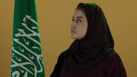 الاحتفال بذكرى اليوم الوطني السعودي، يوم العلم السعودي 11 مارس، مفهوم حب الوطن والنتماء إليه،  بورتريه لامرأة عربية خليجية سعودية محجبة تقف وتحمل علم الوطن بثقة واعتزاز، العيد الوطني السعودي 23 سبتمبر، إظهار الاخلاص والحب للبلاد، خلفية بيج.