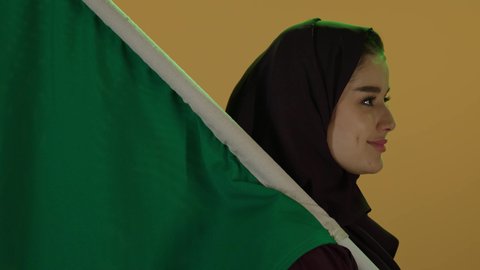 مفهوم حب الوطن والنتماء إليه، يوم العلم السعودي 11 مارس، بورتريه لامرأة عربية خليجية سعودية محجبة تقف وتحمل علم الوطن بثقة واعتزاز، الاحتفال بذكرى اليوم الوطني السعودي، العيد الوطني السعودي 23 سبتمبر، إظهار الاخلاص والحب للبلاد، خلفية بيج.