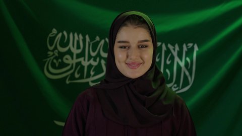 Gestures of pride and dignity, showing loyalty and love for the country, celebrating the anniversary of Saudi National Day, Saudi Flag Day, March 11, the concept of love for the homeland and belonging to it, a portrait of a veiled Saudi Arabian Gulf woman standing in front of the homeland's flag with confidence and pride, beige background.