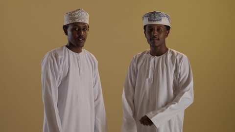 الوقوف بايدى متقاطعة, بورتريه لتوأمان ذو بشرة سمراء عربيان خليجيان عمانيان يرتديان الدشداشة والكمه ينظران الى الكاميرا، إيماءات وجه ويدين تدل على السعادة الثقة، خلفية بنية