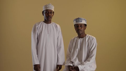 إيماءات وجه ويدين تدل على السعادة والثقة, بورتريه لتوأمان ذو بشرة سمراء عربيان خليجيان عمانيان يرتديان الدشداشة والكمه ينظران الى الكاميرا بتعابير السرور و الفرح، اخ توأم يضع يديه على كتف اخيه، خلفية بنية