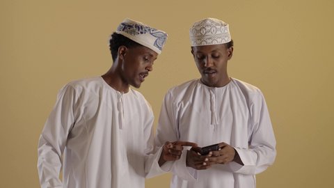 مفهوم التسوق الالكتروني، شراء الهدايا والمستلزمات، الاستفادة من العروض والخصومات, بورتريه لتوأمان ذو بشرة سمراء عربيان خليجيان عمانيان يرتديان الدشداشة والكمه يحملان اكياس ملونة، خلفية بنية