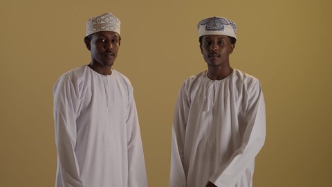 الوقوف باستقامة وأيدي متقاطعة، تعابير وايماءات الثقة بالنفس، بورتريه لتوأمان ذو بشرة سمراء عربيان خليجيان عمانيان يرتديان الدشداشة والكمه ينظران الى الكاميرا، خلفية بنية