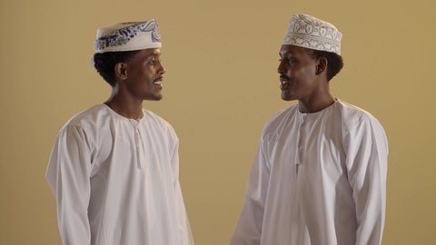 شراء الهدايا والمستلزمات، الاستفادة من العروض والخصومات, مفهوم التسوق الالكتروني، بورتريه لتوأمان ذو بشرة سمراء عربيان خليجيان عمانيان يرتديان الدشداشة والكمه يحملان اكياس ملونة، خلفية بنية