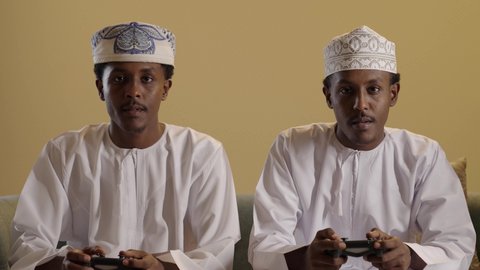بورتريه مقرب لتوأمان ذو بشرة سمراء عربيان خليجيان عمانيان يرتديان الدشداشة والكمه يلعبان لعبة فيديو يحمل كل منهما عصا التحكم، استخدام الأجهزة التقنية الحديثة، ايماءات الانسجام والاستمتاع بالالعاب الالكترونيه، خلفية بنية