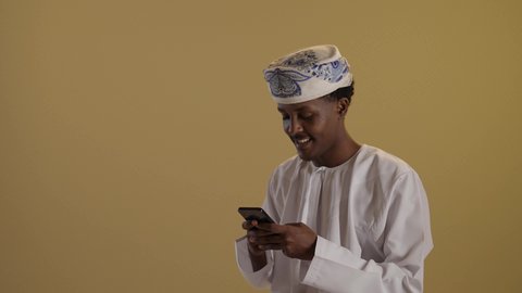 التسوق عبر الانترنت، بورتريه مقرب لتوأمان ذو بشرة سمراء عربيان خليجيان عمانيان يرتديان الدشداشة والكمه يقومان بتصفح الهاتف المحمول، استخدام الأجهزة التقنية الحديثة، دمج التقنية والتكنولوجيا في الحياة اليومية، التواصل مع الأهل والاصدقاء، خلفية بنية