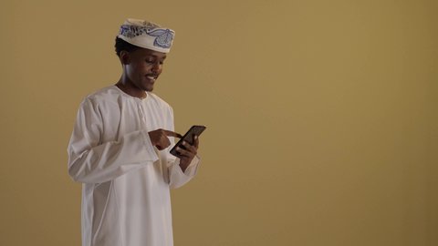 التواصل مع الأهل والاصدقاء، بورتريه لتوأمان ذو بشرة سمراء عربيان خليجيان عمانيان يرتديان الدشداشة والكمه يقومان بتصفح الهاتف المحمول، استخدام الأجهزة التقنية الحديثة، دمج التقنية والتكنولوجيا في الحياة اليومية، التواصل مع الأهل والاصدقاء، خلفية بنية