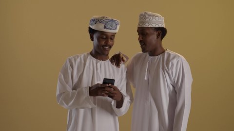 الدردشة والتواصل مع الاصدقاء، بورتريه مقرب لتوأمان ذو بشرة سمراء عربيان خليجيان عمانيان يرتديان الدشداشة والكمه يقومان بتصفح الهاتف المحمول، استخدام الأجهزة التقنية الحديثة، دمج التقنية والتكنولوجيا في الحياة اليومية، التواصل مع الأهل والاصدقاء، خلفية بنية