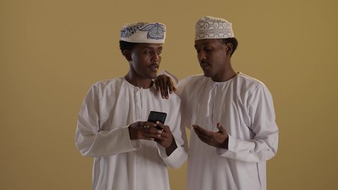 استخدام الأجهزة التقنية الحديثة، دمج التقنية والتكنولوجيا في الحياة اليومية، التواصل مع الأهل والاصدقاء، تصفح مواقع التواصل الاجتماعى، بورتريه لتوأمان ذو بشرة سمراء عربيان خليجيان عمانيان يرتديان الدشداشة والكمه يقومان باستخدام الموبايل، خلفية بنية