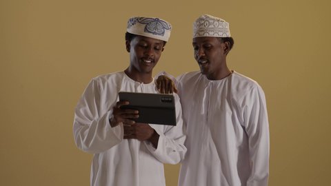 قضاء وقت ممتع على الإنترنت، استخدام الأجهزة التقنية الحديثة والمتطورة في المجال الدراسي، اللعب على الجهاز اللوحي، بورتريه لتوأمان ذو بشرة سمراء عربيان خليجيان عمانيان يرتديان الدشداشة والكمه يستخدمان التابلت، خلفية بنية