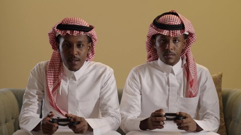 Gestures of harmony and enjoyment in electronic video games, a portrait of twin Arab Gulf Saudis with brown skin wearing the Saudi thobe and ghutrah playing a video game, each holding a controller, with a brown background.