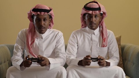 بورتريه لتوأم ذو بشرة سمراء عربيان خليجيان سعوديان يرتديان الثوب والشماغ السعودي يلعبان لعبة فيديو يحمل كل منهما عصا التحكم، ايماءات الانسجام والاستمتاع بالالعاب الالكترونيه، خلفية بنية