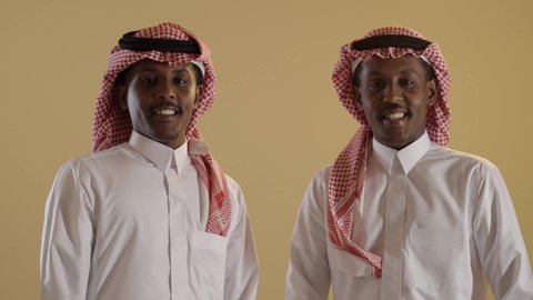 A portrait of twin students with brown skin, Arab, Gulf, and Saudi, wearing the Saudi thobe and shemagh, standing upright, with arms crossed, standing confidently, facial and hand gestures indicating happiness and confidence, with a brown background.