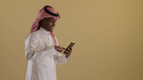 دمج التقنية والتكنولوجيا في الحياة اليومية، التواصل مع الأهل والاصدقاء، تصفح مواقع التواصل الاجتماعى، بورتريه لطالبان توأم ذو بشرة سمراء عربيان خليجيان سعوديان يرتديان الثوب والشماغ السعودي يقومان باستخدام الموبايل، خلفية بنية