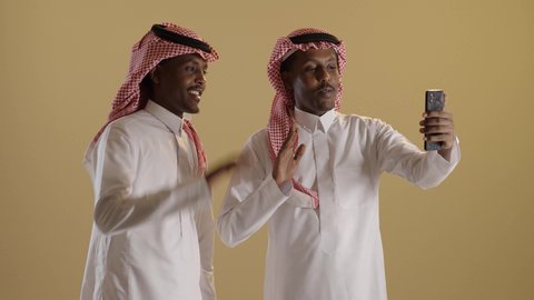 رفع الكف للأعلى بايماءات التحية والسلام، استخدام الأجهزة التقنية الحديثة، التواصل مع الأهل والاصدقاء، اجراء مكالمة فيديوعبرالهاتف المحمول، بورتريه لطالبان توأم ذو بشرة سمراء عربيان خليجيان سعوديان يرتديان الثوب والشماغ السعودي يقومان باستخدام الجوال، خلفية بنية