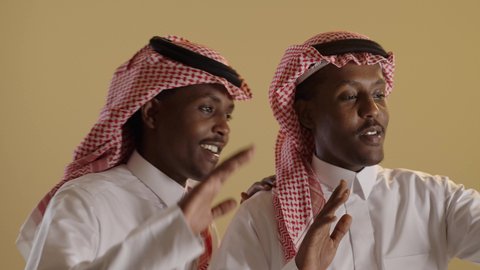 اجراء مكالمة الفيديو باستخدام الهاتف المحمول، الدردشة والتواصل مع الاصدقاء، بورتريه مقرب لطالبان توأم ذو بشرة سمراء عربيان خليجيان سعوديان يرتديان الثوب والشماغ السعودي، خلفية بنية