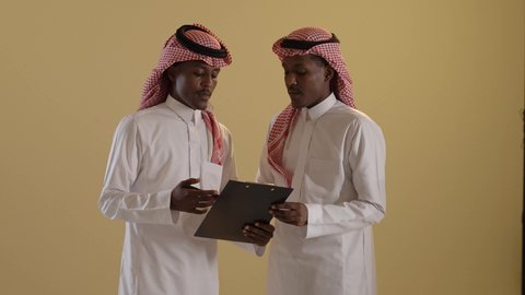 المشاركة والتعاون، نتائج بحث ودراسة موضوع، بورتريه مقرب لطالبان توأم ذو بشرة سمراء عربيان خليجيان سعوديان يرتديان الثوب والشماغ السعودي يقومان بتدوين المهام اليومية، استخدام اوراق بيضاء وقلم، خلفية بنية