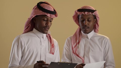 Monitoring plans and projects, research results and study of a topic, a close portrait of twin students with brown skin, Arab Gulf Saudis wearing the Saudi thobe and shemagh, writing down daily tasks, using white papers and a pen, brown background.