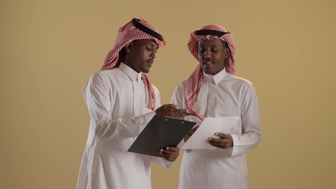 نتائج بحث ودراسة موضوع، بورتريه مقرب لطالبان توأم ذو بشرة سمراء عربيان خليجيان سعوديان يرتديان الثوب والشماغ السعودي يقومان بتدوين المهام اليومية، استخدام اوراق بيضاء وقلم، خلفية بنية