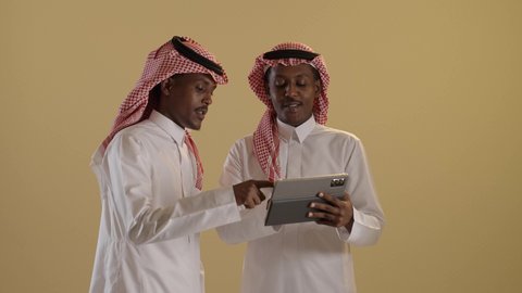 قضاء وقت ممتع على الإنترنت، استخدام الأجهزة التقنية الحديثة والمتطورة في المجال الدراسي، اللعب على الجهاز اللوحي، بورتريه مقرب لطالبان توأم ذو بشرة سمراء عربيان خليجيان سعوديان يرتديان الثوب والشماغ السعودي يستخدمان التابلت، خلفية بنية