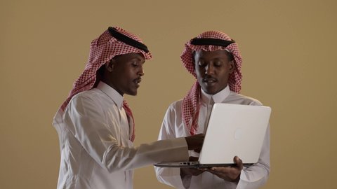 استخدام الاجهزة التقنية الحديث، بورتريه لطالبان توأم ذو بشرة سمراء عربيان خليجيان سعوديان يرتديان الثوب والشماغ السعودي يستخدمان اللابتوب، المذاكرة والدراسة عن بعد، المشاركة والتعاون، خلفية بنية