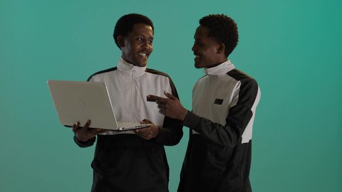 The use of modern technological devices, a portrait of twin students with brown skin, Arab Gulf Saudi nationals wearing school uniforms, using a laptop, participation and collaboration, studying and remote learning, green background.