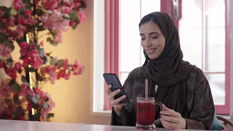 الاستمتاع بالمشروبات الباردة في فصل الصيف, الاستمتاع بالأنشطة الترفيهية, امرأة عربية خليجية سعودية محجبة ترتدي عباءة تجلس في كافي شوب راقي تقوم باستخدام الهاتف الجوال، إيماءات السعادة و السرور, قضاء الأوقات الممتعة في الخارج