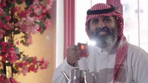 أجواء الهدوء والاسترخاء والراحة، تصوير مقرب لرجل خليجي عربي سعودي يرتدي الثوب التقليدي والشماغ يجلس على الكرسي يمسك بيده كوب ينظر إلى الكاميرا، الاستمتاع بشرب المشروبات الساخنة، إكرام الضيف وحسن الاستقبال والضيافة، مفهوم الرجولة والاناقة