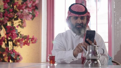 الاستمتاع بشرب المشروبات الساخنة، أجواء الهدوء والاسترخاء والراحة داخل كافيه راقي، تصوير مقرب لرجل خليجي عربي سعودي يرتدي الثوب التقليدي والشماغ يجلس على الكرسي يمسك بيده هاتف محمول وبجانبه كأس من الشاي، إكرام الضيف وحسن الاستقبال والضيافة،مفهوم الرجولة والاناقة