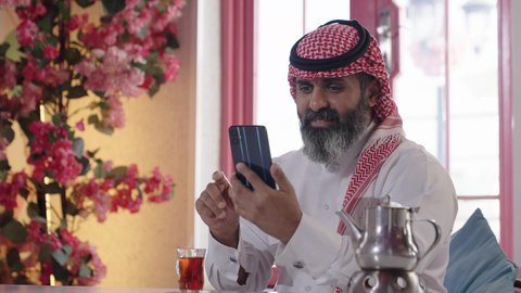إكرام الضيف وحسن الاستقبال والضيافة، الاستمتاع بشرب المشروبات الساخنة، أجواء الهدوء والاسترخاء والراحة داخل كافيه راقي، تصوير مقرب لرجل خليجي عربي سعودي يرتدي الثوب التقليدي والشماغ يجلس على الكرسي يمسك بيده هاتف محمول وبجانبه كأس من الشاي، مفهوم الرجولة والاناقة