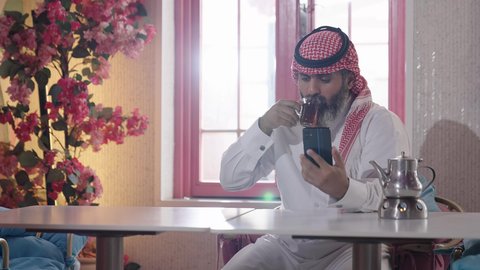 شرب الشاي الاحمر، إكرام الضيف وحسن الاستقبال والضيافة، الاستمتاع بشرب المشروبات الساخنة، أجواء الهدوء والاسترخاء والراحة داخل كافيه راقي، تصوير مقرب لرجل خليجي عربي سعودي يرتدي الثوب التقليدي والشماغ يجلس على الكرسي يمسك بيده هاتف محمول وبجانبه كأس من الشاي
