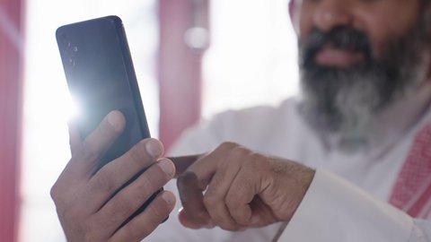 تصفح مواقع التواصل الاجتماعي، إكرام الضيف وحسن الاستقبال والضيافة، الاستمتاع بشرب المشروبات الساخنة، أجواء الهدوء والاسترخاء والراحة داخل كافيه راقي، تصوير مقرب لرجل خليجي عربي سعودي يرتدي الثوب التقليدي والشماغ يجلس على الكرسي يمسك بيده هاتف محمول