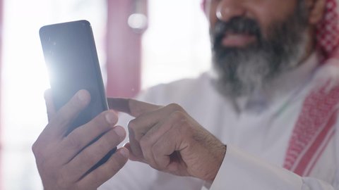 تقليب وتصفح مواقع التواصل الاجتماعي، إكرام الضيف وحسن الاستقبال والضيافة، الاستمتاع بشرب المشروبات الساخنة، أجواء الهدوء والاسترخاء والراحة داخل كافيه راقي، تصوير مقرب لرجل خليجي عربي سعودي يرتدي الثوب التقليدي والشماغ يجلس على الكرسي يمسك بيده هاتف محمول