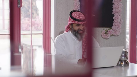 انهاء مهام العمل عن بعد، إكرام الضيف وحسن الاستقبال والضيافة، الاستمتاع بشرب المشروبات الساخنة، أجواء الهدوء والاسترخاء والراحة داخل كافيه راقي، تصوير مقرب لرجل خليجي عربي سعودي يرتدي الثوب التقليدي والشماغ يجلس على الكرسي يعمل على حاسب محمول وبجانبه كأس من الشاي