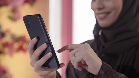 A Saudi woman is making a video call via her mobile phone, integrating advanced technology into daily life, communicating with family and friends. A Gulf Arab Saudi woman wearing a hijab and an abaya sits on a chair in one of the upscale cafes, enjoying moments of tranquility and relaxation.