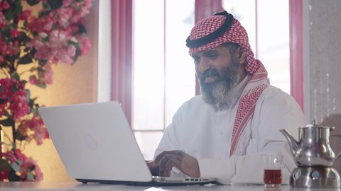Looking at the camera with gestures of pleasure, honoring the guest and showing warm hospitality, enjoying hot drinks, the atmosphere of tranquility and relaxation inside an upscale cafe, a close-up shot of a Gulf Arab Saudi man wearing the traditional thobe and shemagh, sitting in a chair working on a laptop with a cup of tea beside him