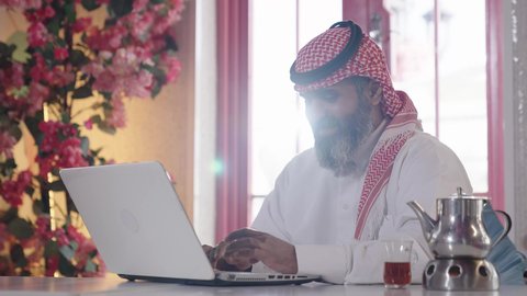 متابعة الأعمال عن بعد، دمج التكنولوجيا مع الحياة اليومية، الاستمتاع بشرب المشروبات الساخنة، أجواء الهدوء والاسترخاء داخل كافيه راقي، تصوير مقرب لرجل خليجي عربي سعودي يرتدي الثوب التقليدي والشماغ يجلس على الكرسي يعمل على حاسب محمول وبجانبه كأس من الشاي